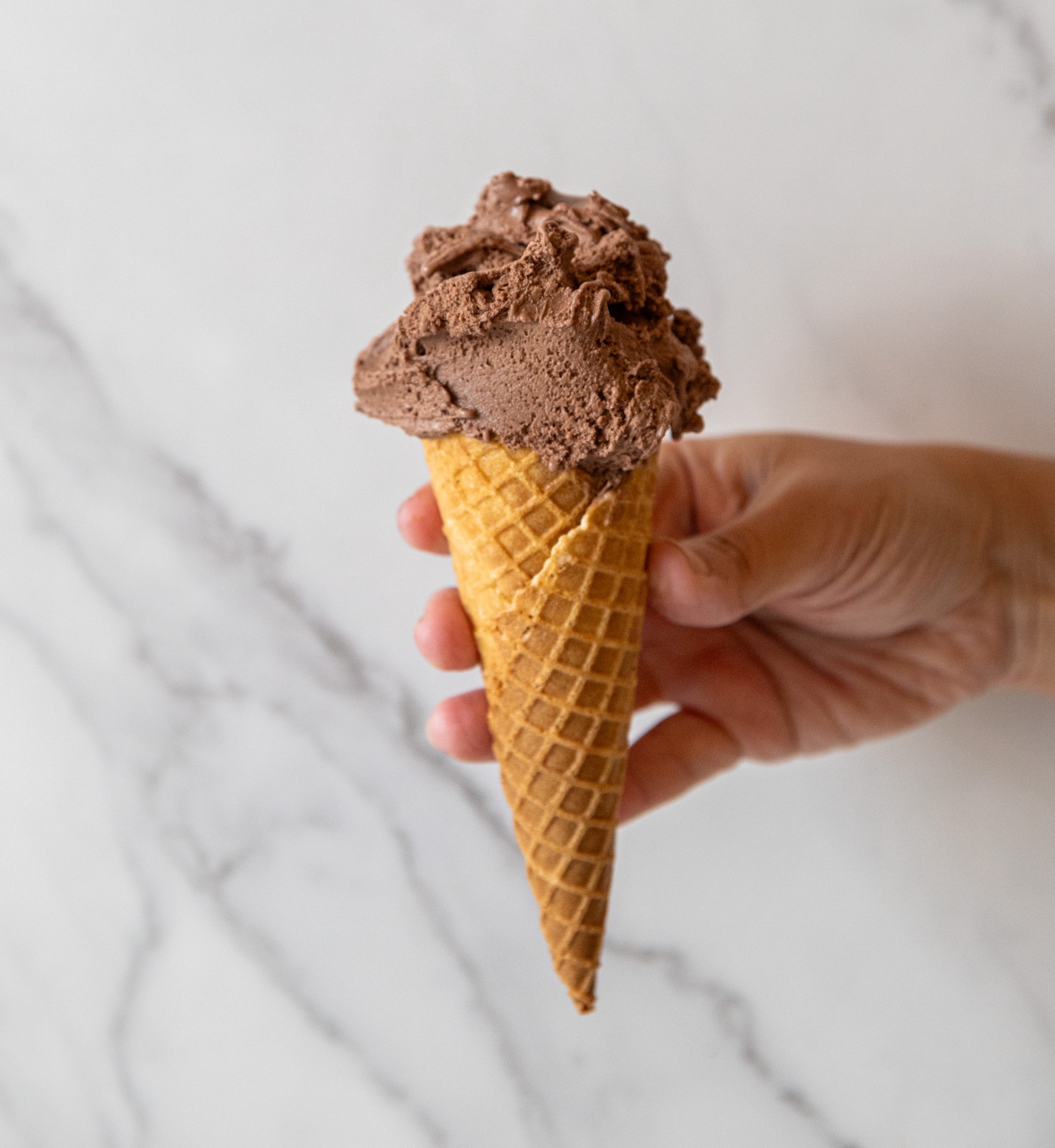 Hand holding a fresh chocolate gelato waffle cone against a marble background